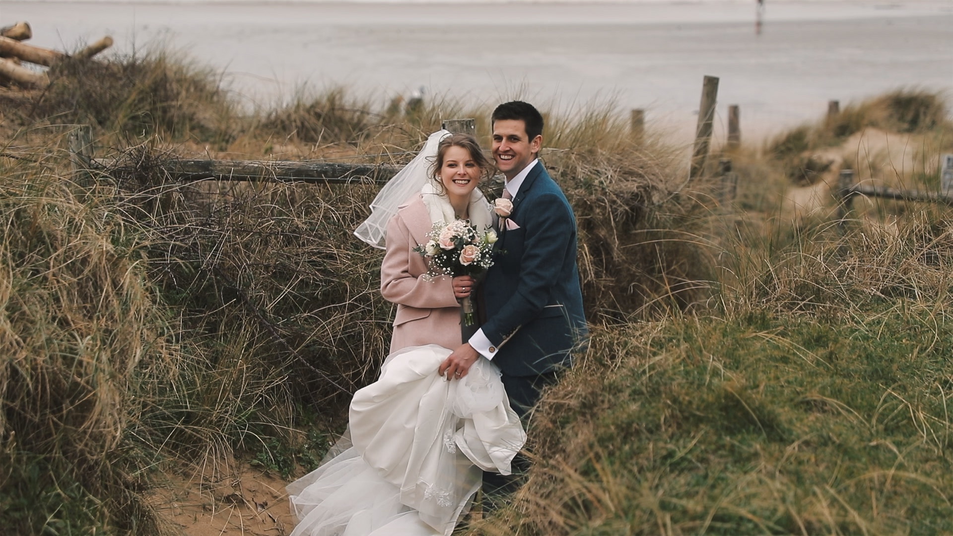 A relaxed coastal wedding moment in Cornwall, where a bride and groom smile on a windswept beach path framed by tall grasses as soft sea air moves through the bride’s veil.