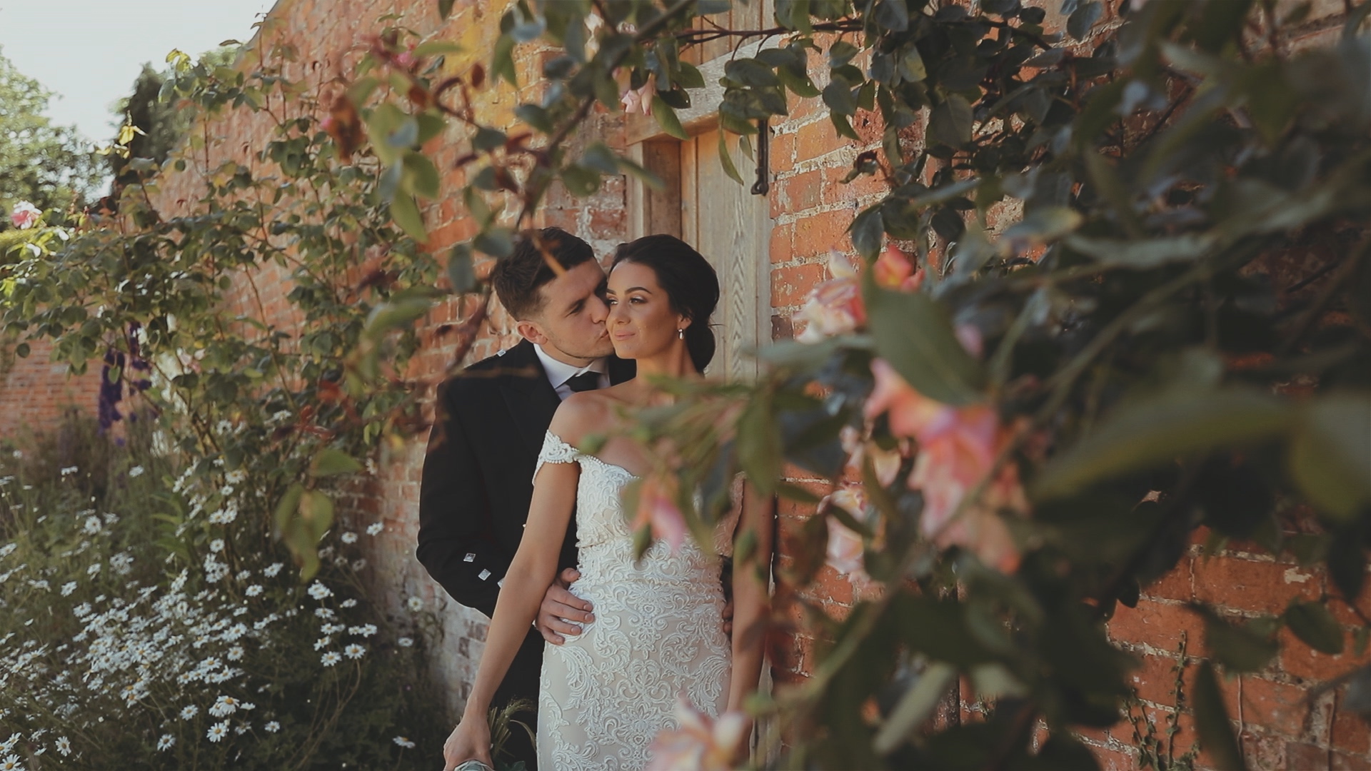 Wedding couple portrait at Elmhay Park in Somerset, embracing by a brick wall with greenery and flowers
