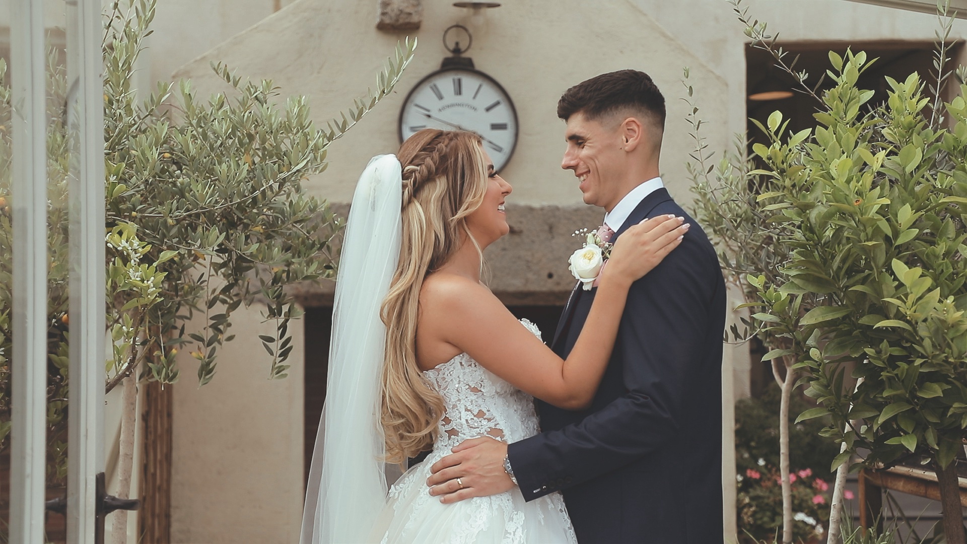 CP-green-house-laugh Bride and groom embrace and smile in front of the greenhouse at Shilstone House in Devon, surrounded by lush garden greenery with a vintage clock detail in the background, capturing a joyful romantic moment.