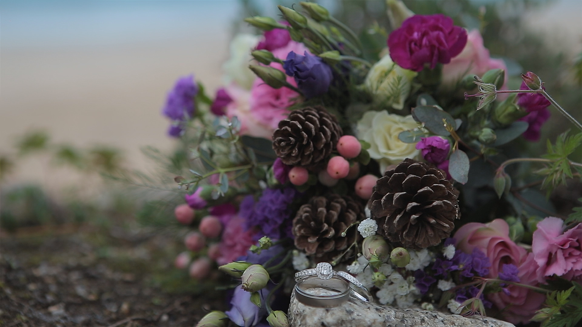 Wedding rings resting beside a vibrant bouquet of pink, purple, and white flowers with pinecones and berries on a stone surface.