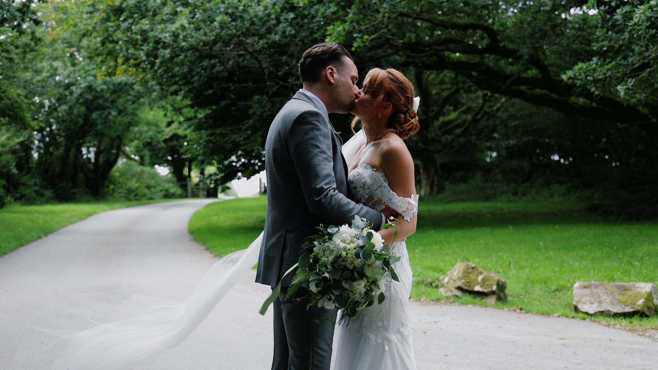 Bride and groom sharing a kiss on a tree-lined path at Trevenna in Cornwall as the bride’s veil flows in the wind