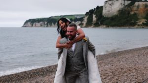 A bride gets a piggy back ride from the groom on Seaton beach captured by South west based wedding videographer