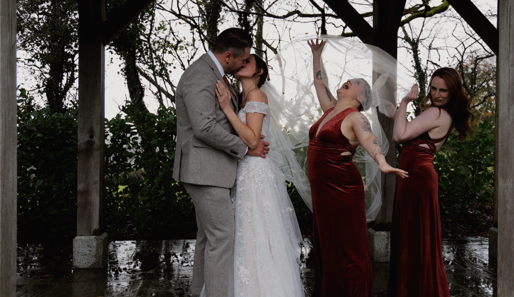 Bride and groom kissing under a wooden gazebo at Trevenna, while bridesmaids in red dresses playfully lift and move the bride’s flowing veil.