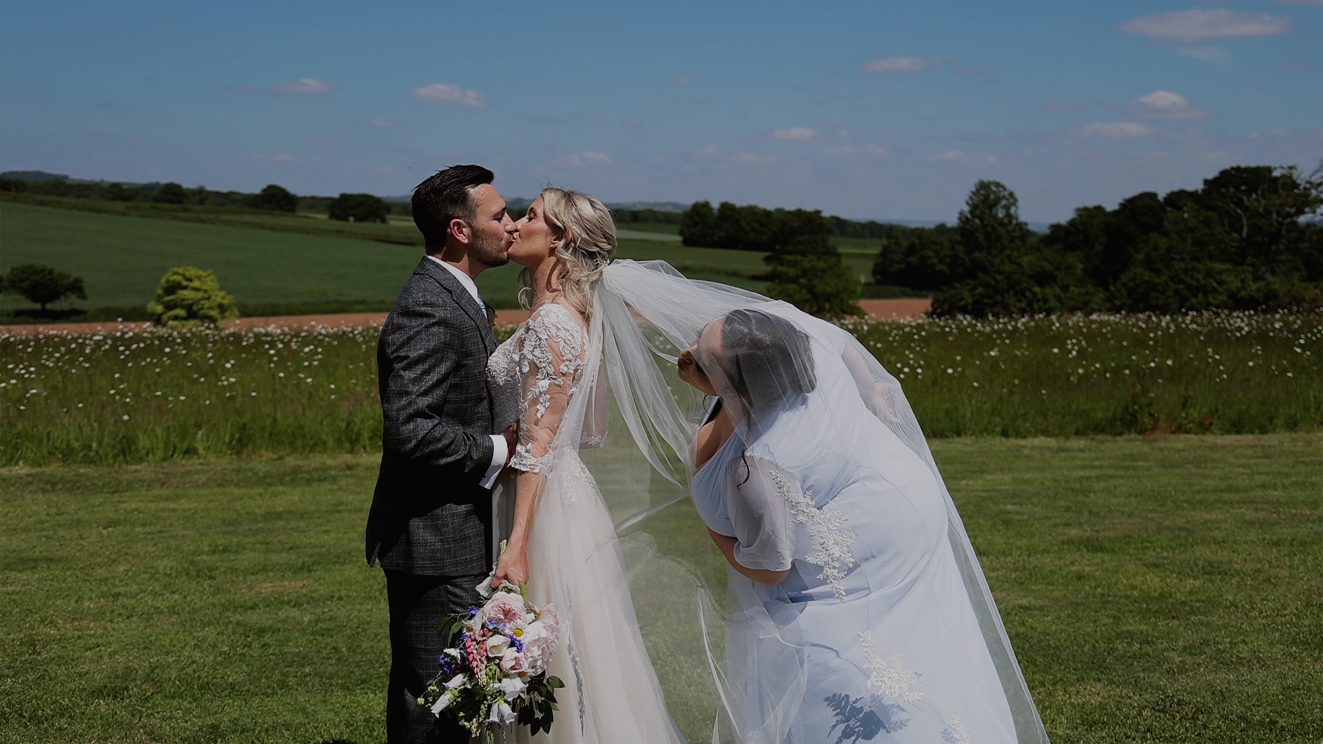 A bridesmaid sneaking under the veil while the couple have a kiss at Harefield Barn