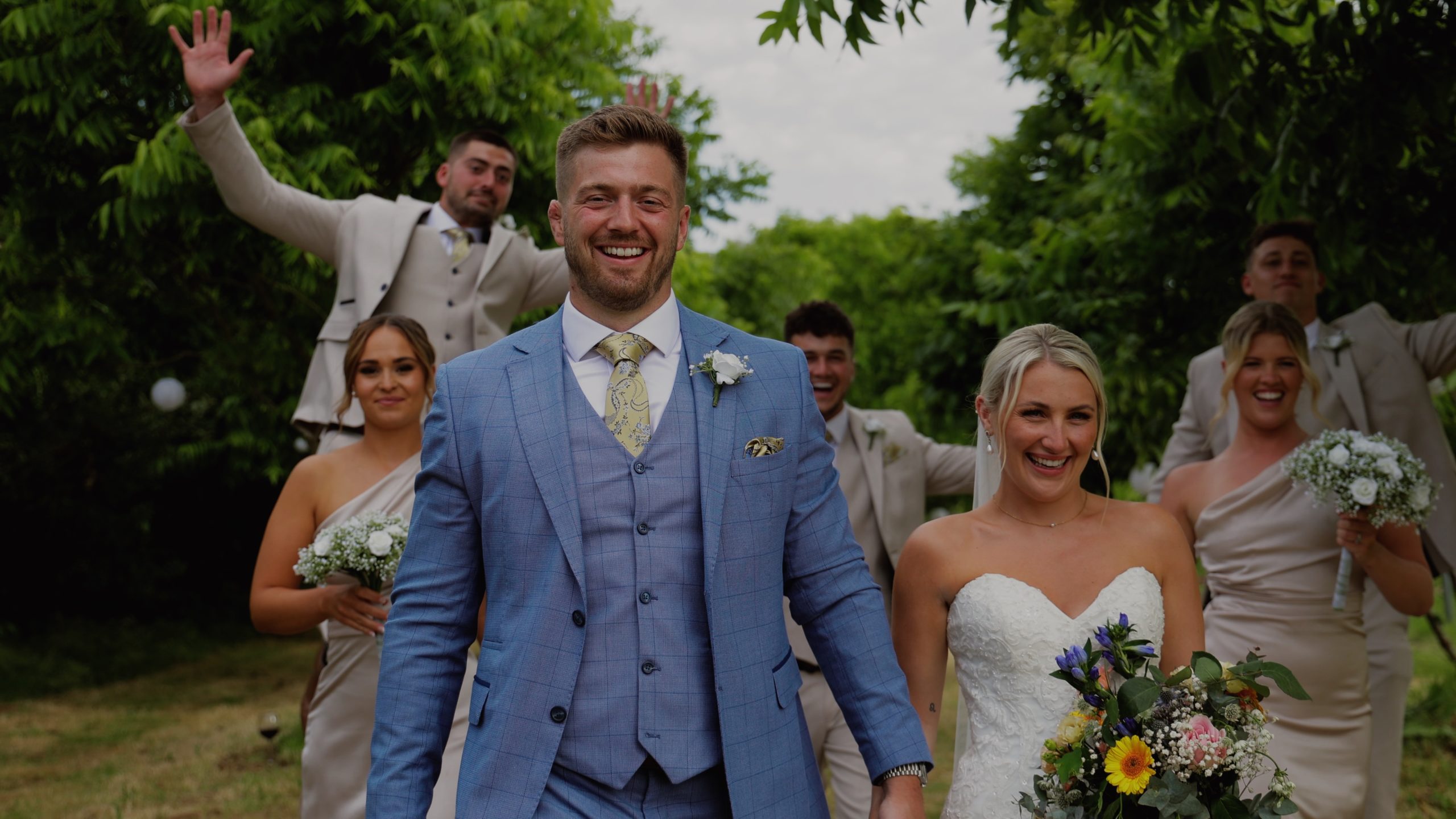 A bride and groom walking along and having fun with their wedding party in the background