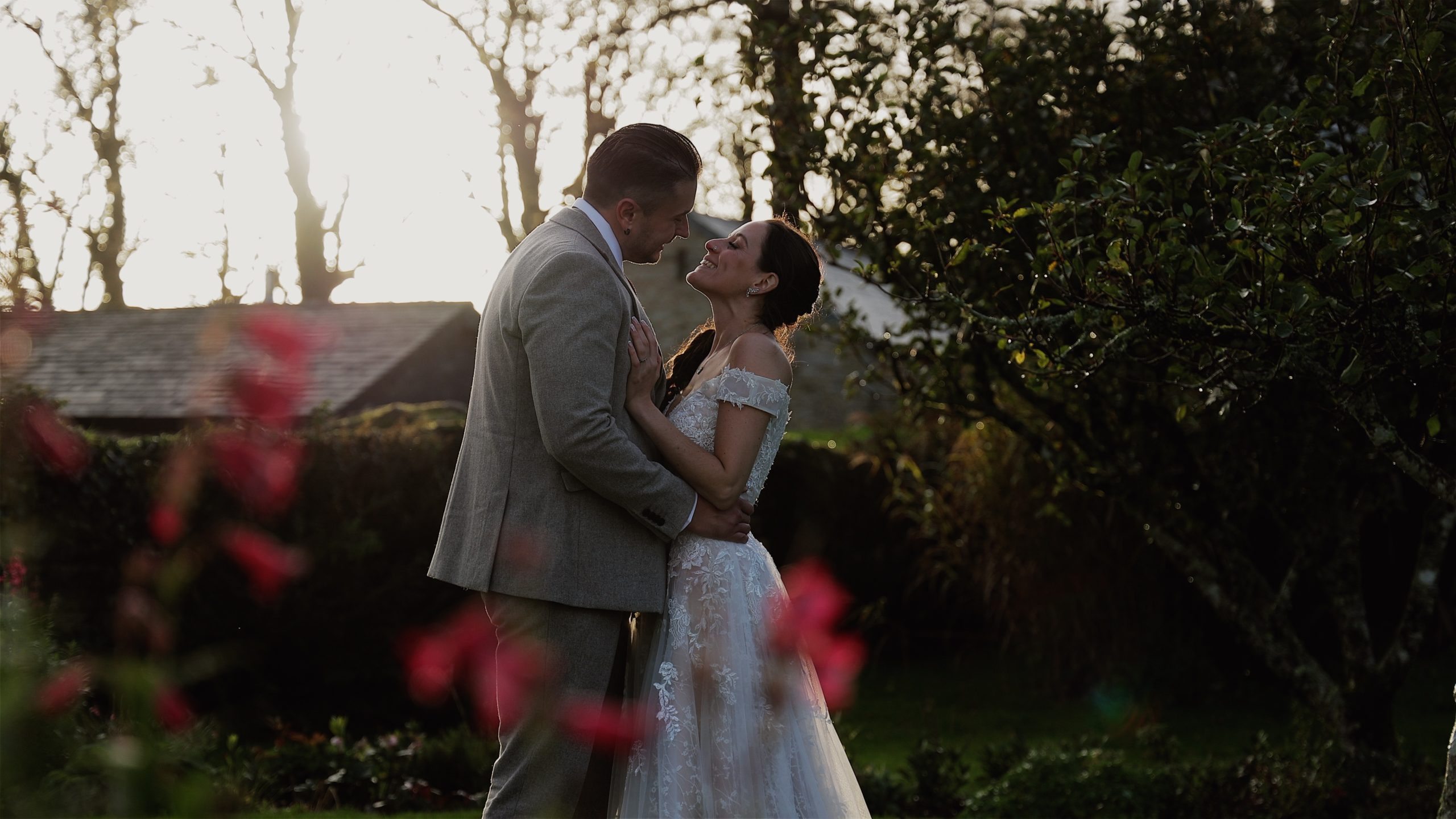 A bride and groom stood in evening sunlight with flowers in the fore ground at Trevenna Barns