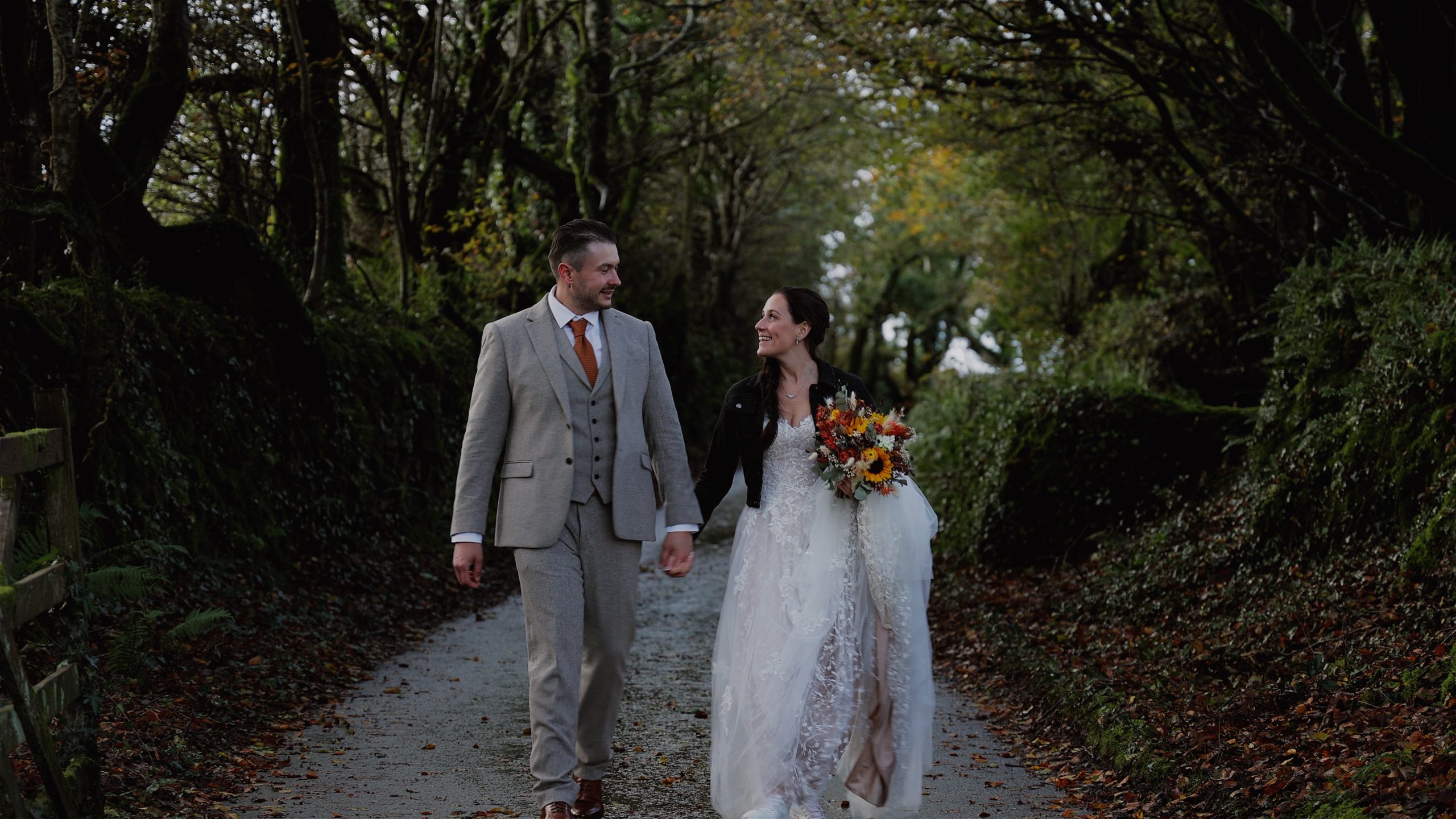 A bride and groom walking through a dark lane covered in trees looking at each other and smiling