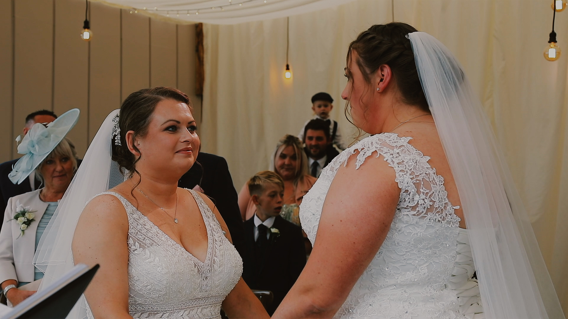 Same sex marriage at Trevenna Barns Two brides in white lace gowns and veils hold hands facing each other during an indoor ceremony at Trevenna Barn in Cornwall, surrounded by smiling guests.