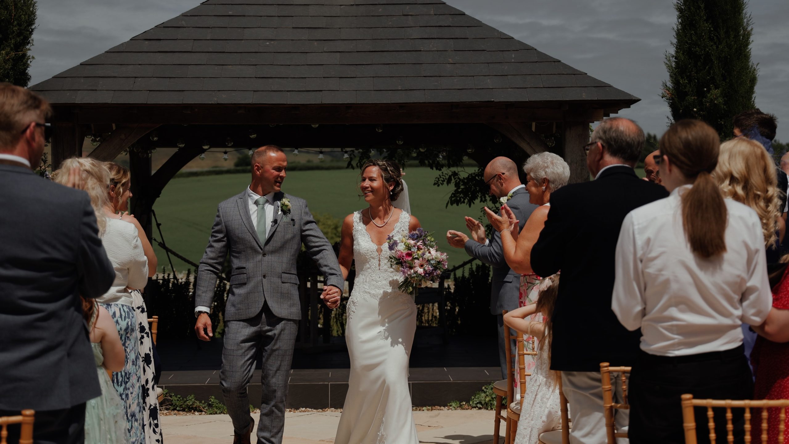 Bride and groom walk hand in hand down an outdoor aisle at Harefield Barn, passing under a wooden gazebo as guests applaud.