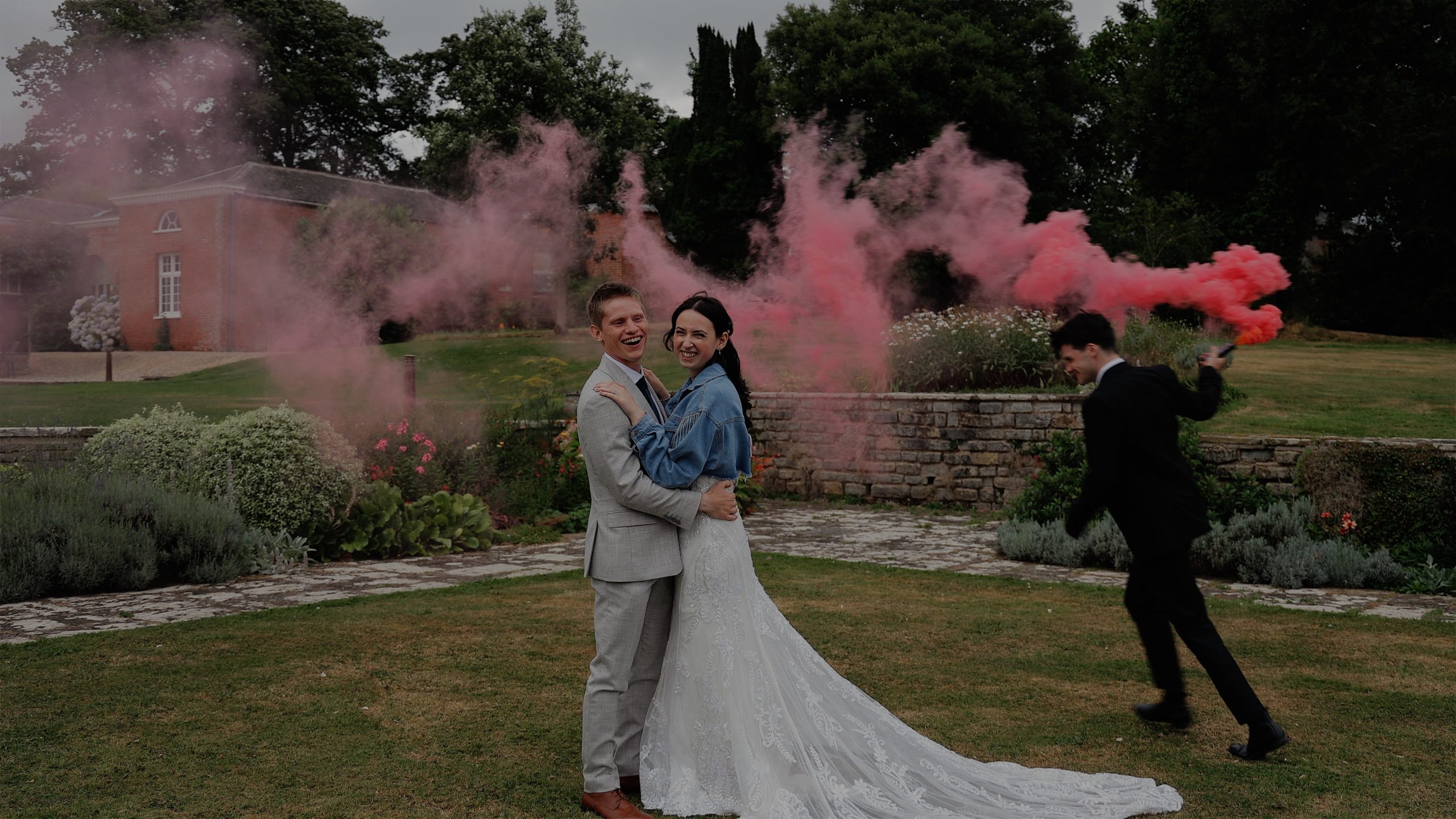 A bride & groom hold each each while their bestman runs around them with a pink smokebomb at Rockbeare Manor