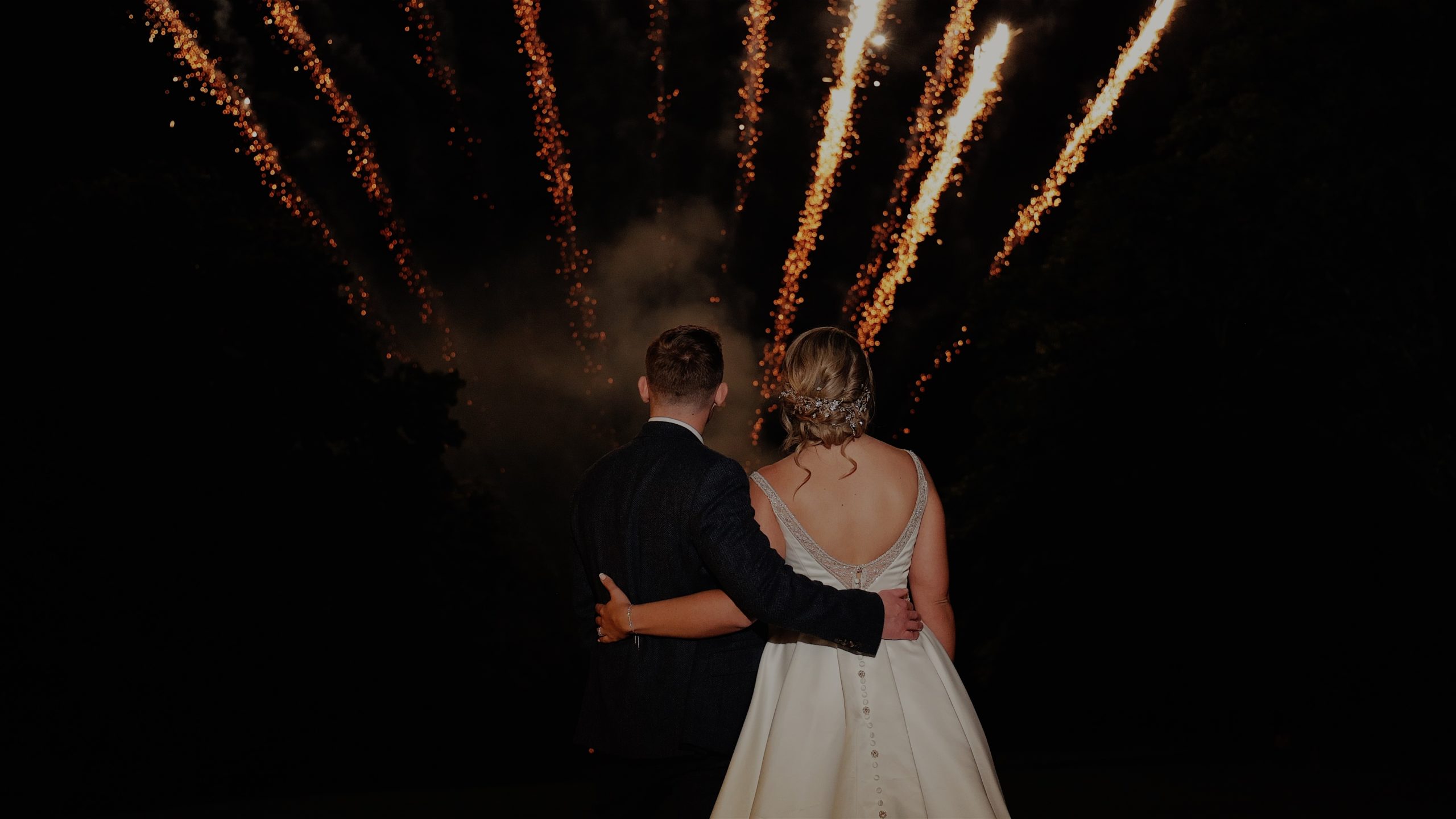 A bride and groom standing with arms around each others backs as they watch fireworks at St Audries Park