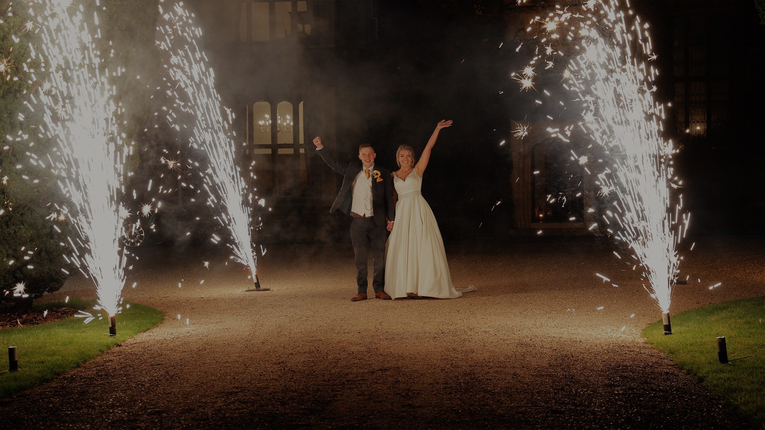 Becca & Andy standing between sparkler towers in front of St Audries Park Newlywed bride & groom celebrate outdoors at night at St Audries Park, surrounded by fireworks in a cinematic wedding moment.