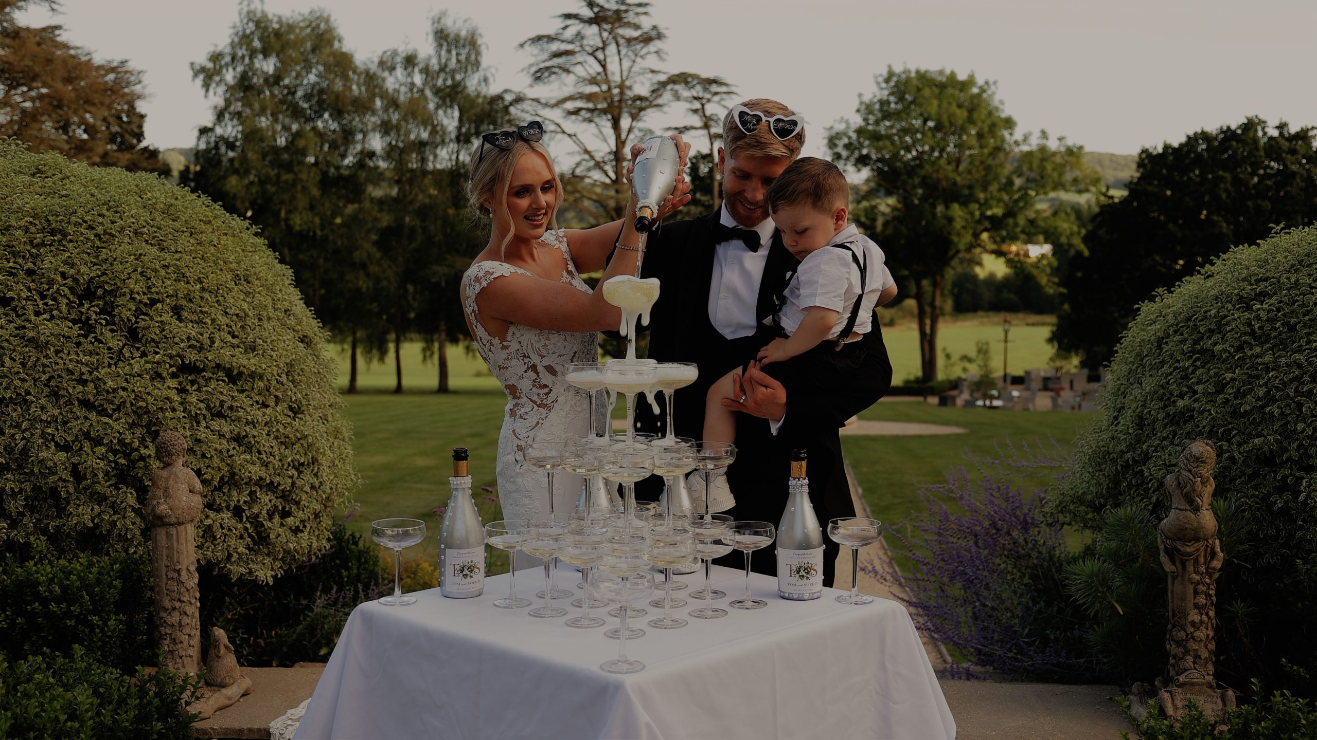 A bride and groom stand in front of a tower of glasses pouring a bottle of champagne at Deer Park country House