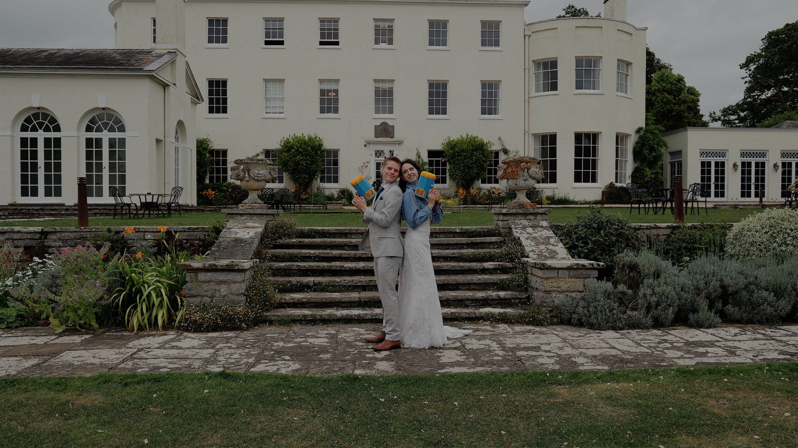 Couple stand back to back holding bubble guns at Rockbeare Manor in Devon, in front of the white historic mansion and manicured gardens.