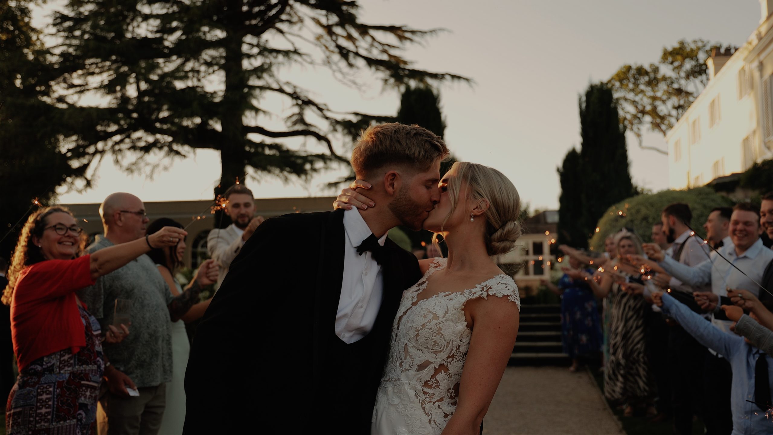 A bride & groom kissing at sunset with sparklers at Deer Park country House Newlywed couple share a kiss on a garden path at Deer Park Country House, surrounded by guests holding sparklers under a twilight sky and tall trees.