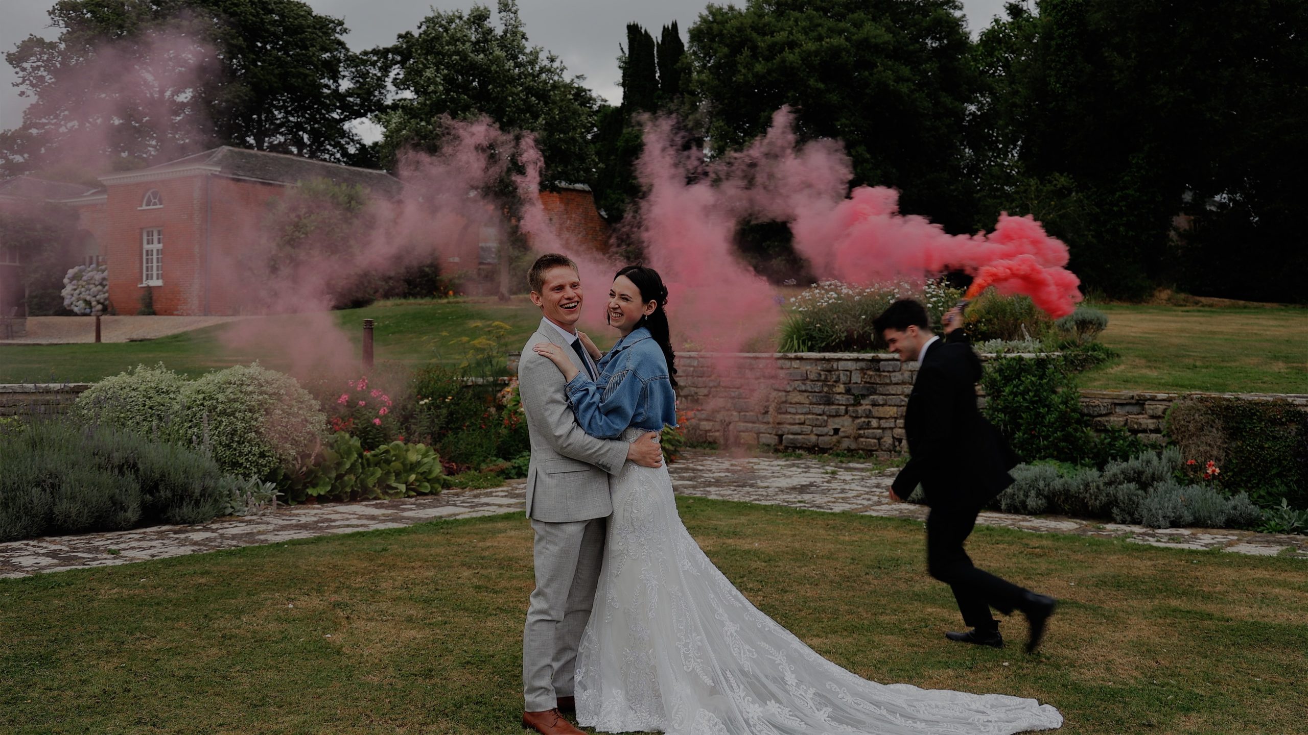 A bride & groom hold each each while their bestman runs around them with a pink smokebomb at Rockbeare Manor