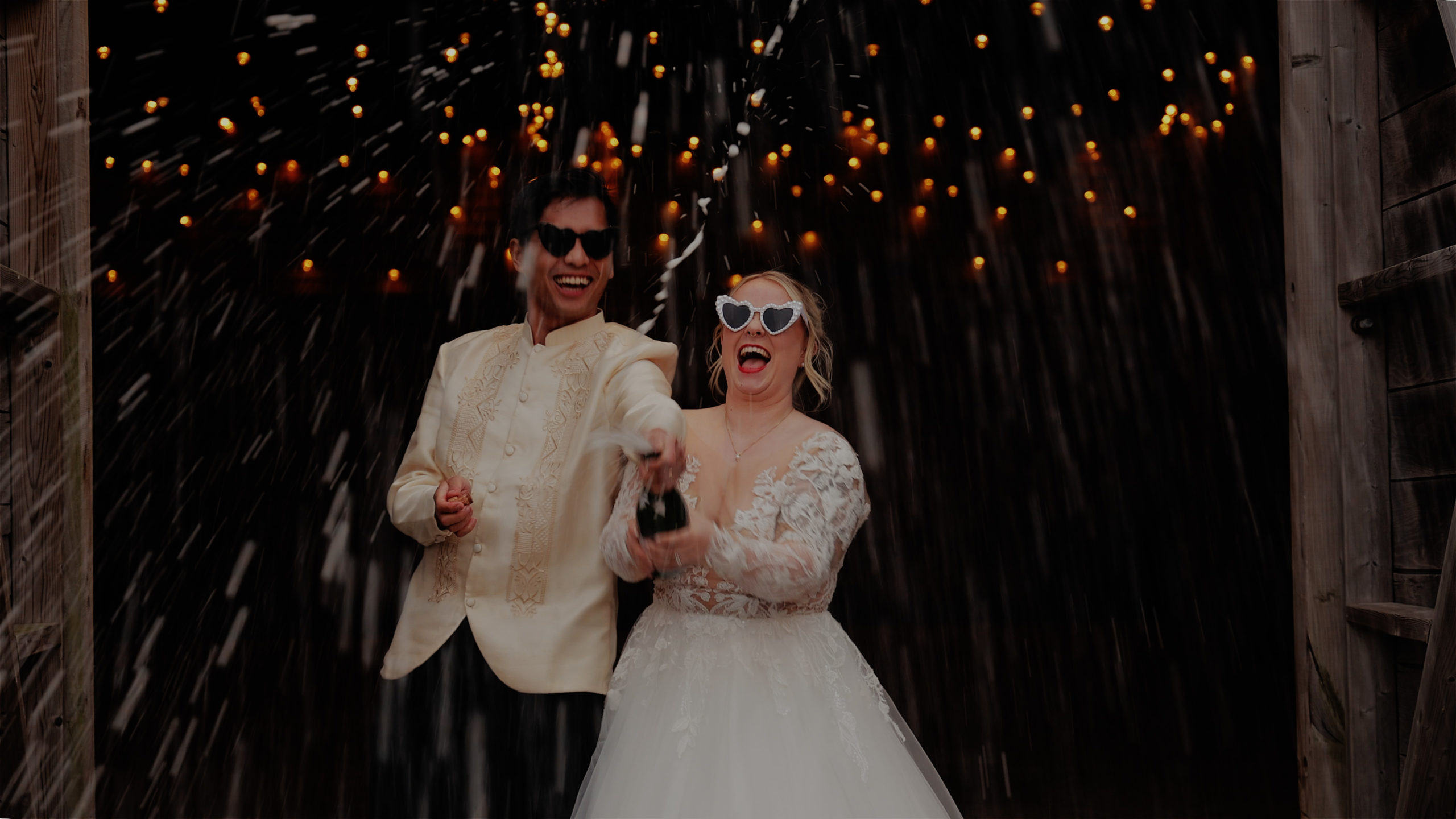 Bride and groom celebrate with champagne in front of a rustic barn at The Green in Cornwall, wearing heart-shaped sunglasses under sparkling string lights.