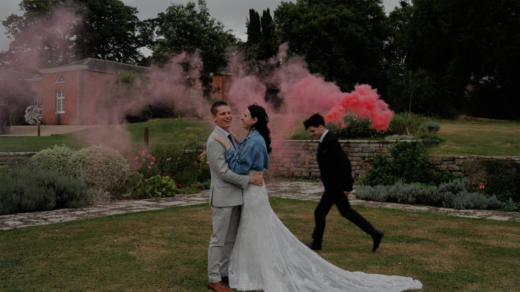 A young wedding couple enjoying a fun moment at Rockbeare Manor, the bride wears a bespoke denim jacket embracing the groom as a groomsman runs around them releasing a pink smoke bomb.