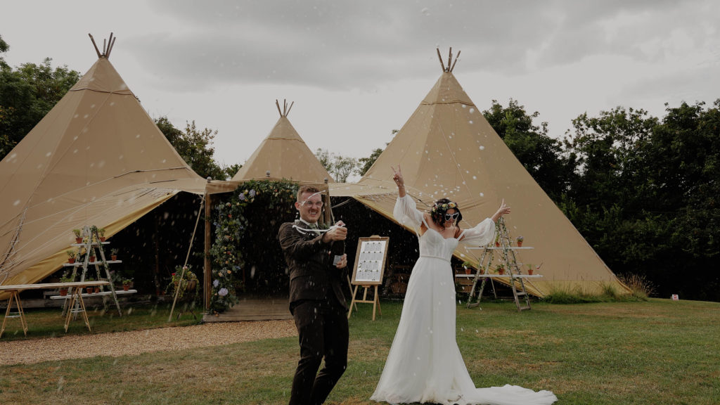 Boho wedding couple celebrating with champagne at Hareston Manor, Plymouth, in front of a tipi tent.