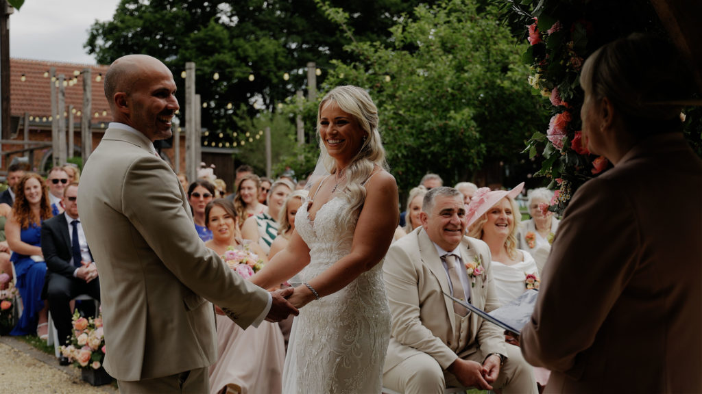 Couple holding hands during their outdoor wedding ceremony under the arbour at Froginwell Vineyard, bride in lace dress, groom in beige suit, joyful guests watching