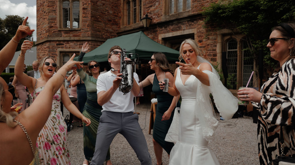 Wedding guests dancing in the gardens with a bride in a white dress while a live saxophonist performs at Mount Edgcumbe House and Country Park, creating a fun outdoor celebration.