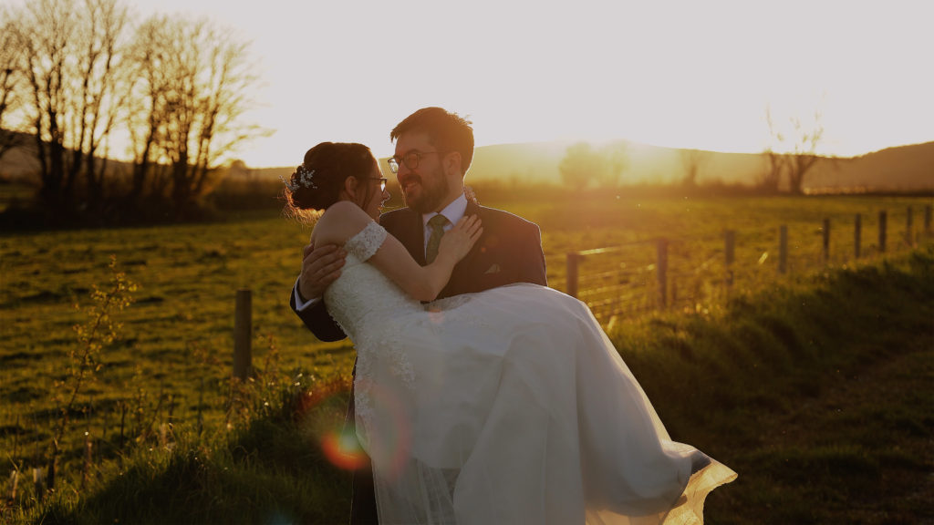 Romantic wedding couple moment with groom carrying bride across a golden sunset field at The Green, surrounded by bare trees and soft evening light.