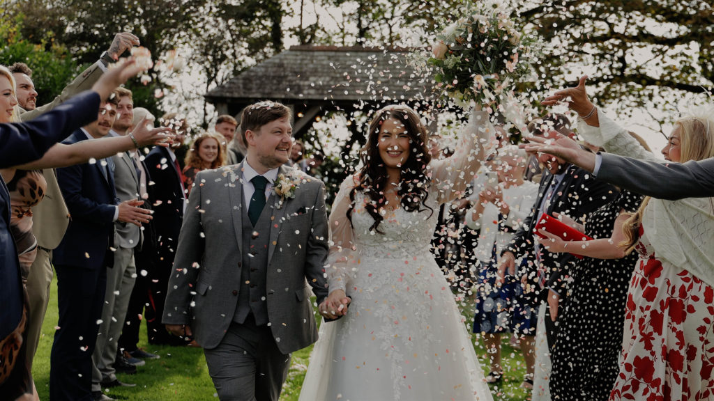 Bride and groom walking through confetti during a garden celebration, with the bride holding her bouquet in the air at Trevenna.