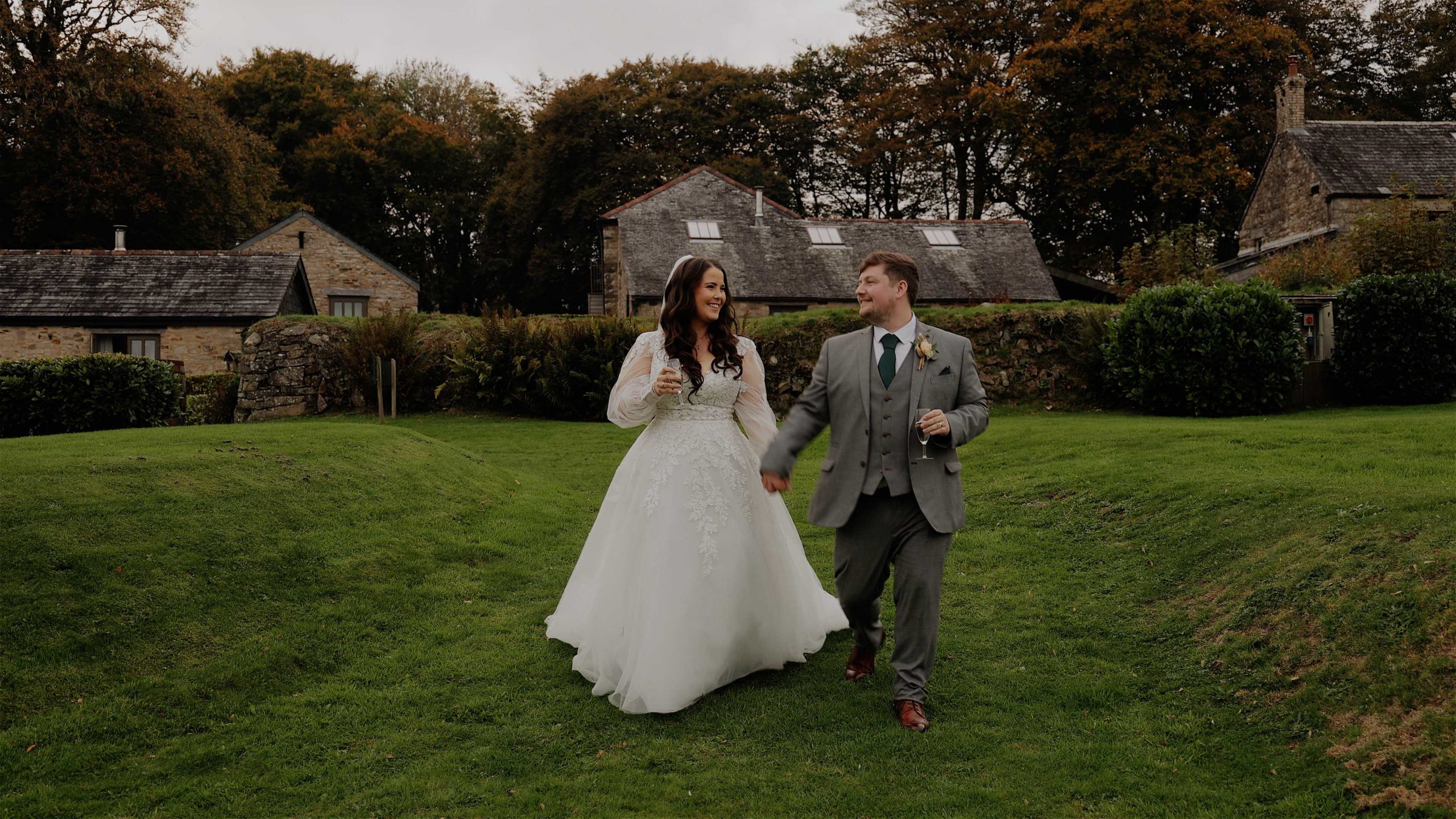 Bride and groom walk hand in hand across a lush green lawn at Trevenna Barn in Cornwall, with rustic stone barns and autumn trees in the background.