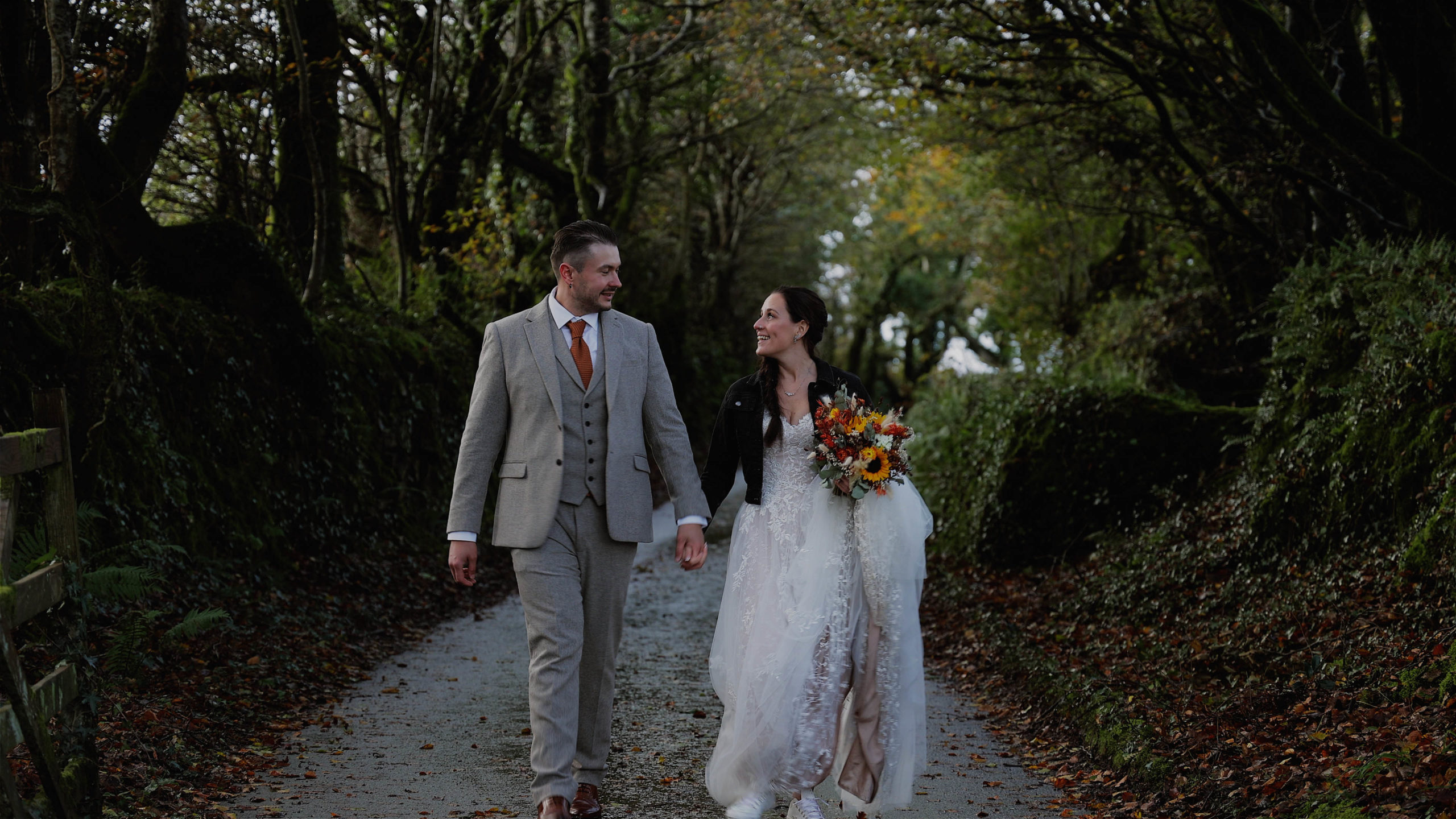 A bride and groom walking through a dark lane covered in trees looking at each other and smiling