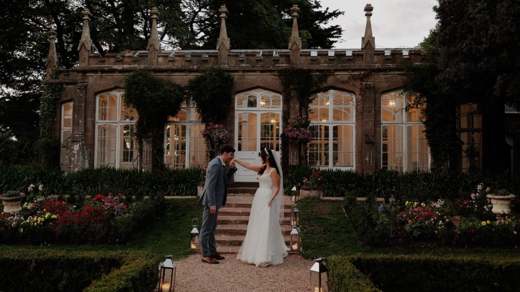 Groom kissing the back of the bride’s hand as they stand in front of a Victorian orangery at St Audries Park, surrounded by elegant gardens.