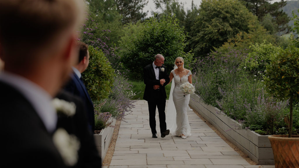 Bride being escorted down an outdoor garden aisle by her father during a wedding ceremony at Deer Park Country House, walking towards the groom as guests watch from the gardens.