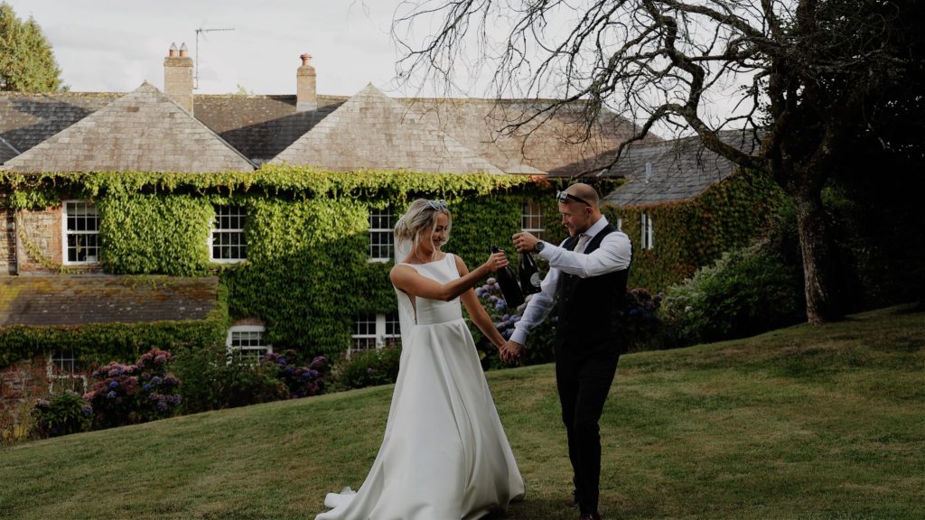 Bride and groom walking across a lawn and clinking champagne bottles together, wearing heart-shaped sunglasses in front of a country house at Wooladon Estate during their wedding day.