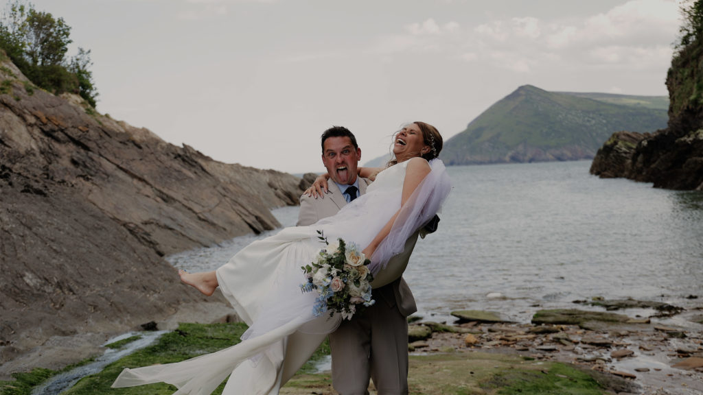 Coastal wedding moment with groom carrying his bride on rocky shoreline at Watermouth Cove, surrounded by cliffs and a calm sea.