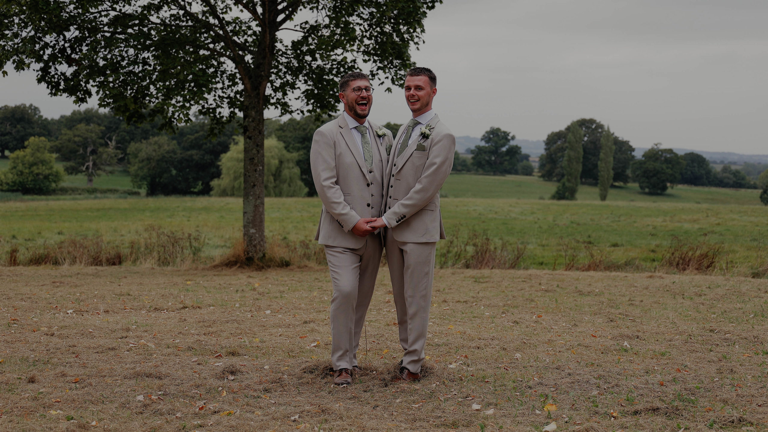 Two grooms in light grey suits share a moment holding hands and laughing in a grassy field at Rockbeare Manor in Devon, beneath a tree during their wedding celebration.