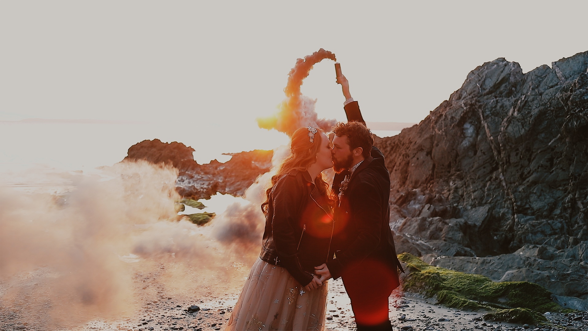 Bride wearing a bespoke leather jacket and groom kissing on a rocky beach at Polhawn Fort, holding a smoke flare at sunset with a warm golden glow.